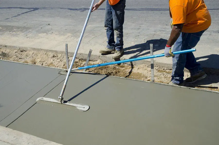Two workers leveling a fresh concrete slab with long screed poles
